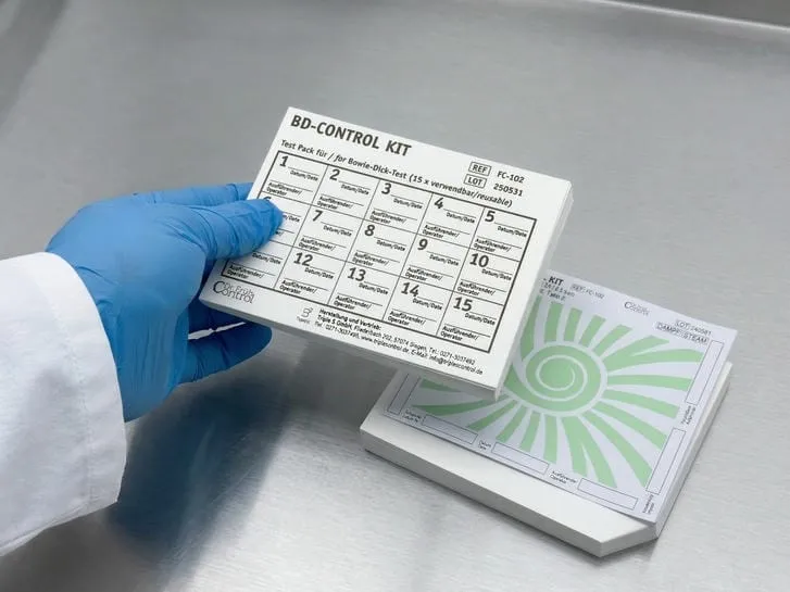 Eine behandschuhte Hand hält ein BD-Control Kit Testpaket mit Feldern für Datum und Ausführenden, daneben liegt ein Indikatorbogen
